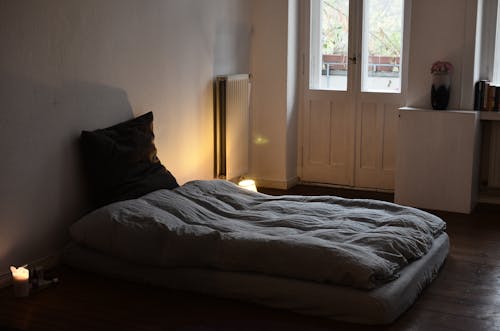 Comfortable mattress with blanket and cushion placed on wooden floor in dark bedroom with burning candles and flowers in vase