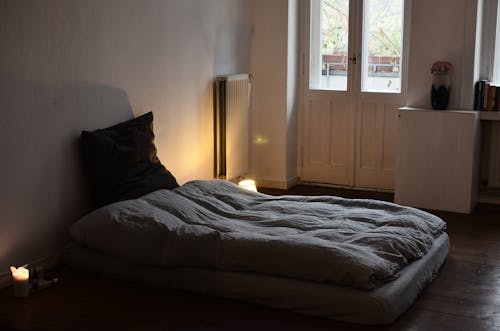 Comfortable mattress with blanket and cushion placed on wooden floor in dark bedroom with burning candles and flowers in vase