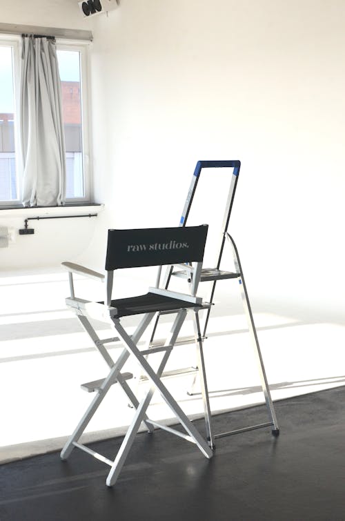 Director chair with inscription and ladder placed in spacious empty studio against big window with curtain in room with white wall