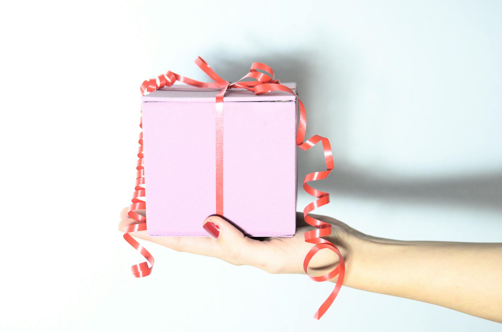 A beautifully wrapped pink gift box with a red ribbon held in a woman's hand, ideal for celebrations.