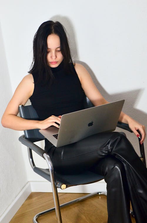 Concentrated female with dark hair sitting on chair with crossed legs and typing on netbook near white wall in corner