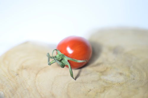 Free Closeup single small ripe cherry tomato placed on shaped wooden cutting board on white table on blurred background in light kitchen Stock Photo