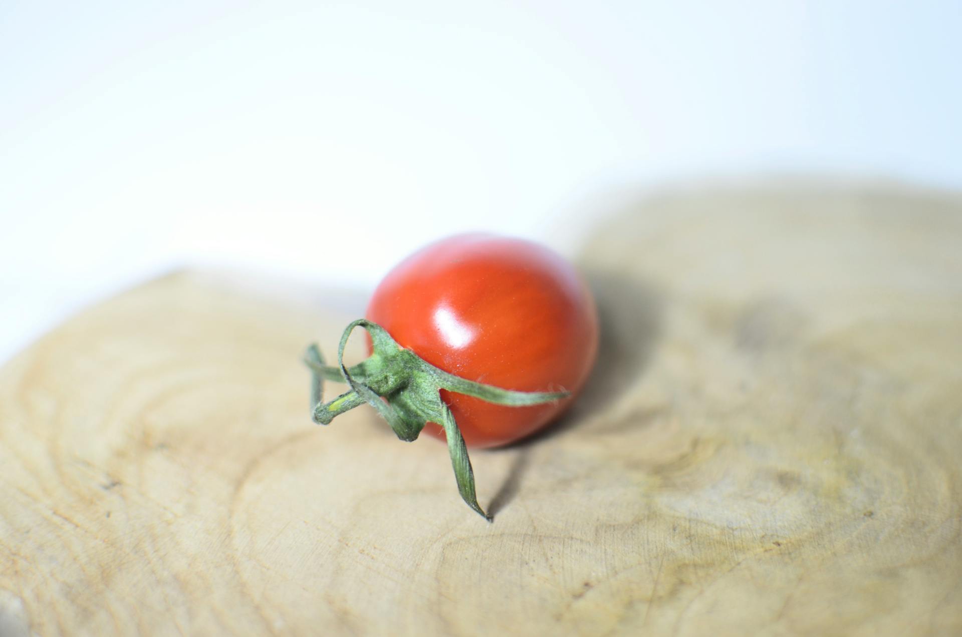 Closeup single small ripe cherry tomato placed on shaped wooden cutting board on white table on blurred background in light kitchen