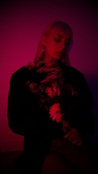 Young pensive female with closed eyes in black wear with flowers standing against wall in dark room illuminated with purple neon light