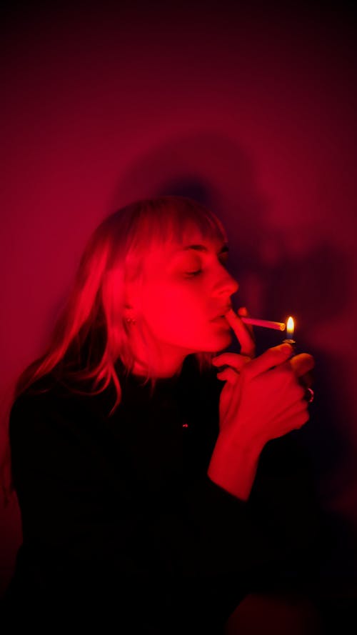 Focused young woman in black outfit lighting cigarette for smoking in dark room illuminated with red neon light