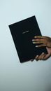 Crop anonymous informal female with tattoos and colorful manicure demonstrating book with black cover against gray background