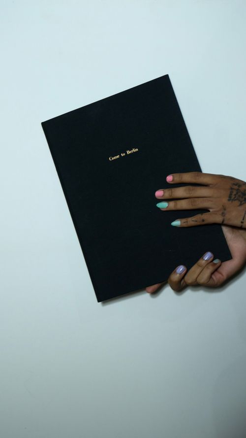 Free Crop anonymous informal female with tattoos and colorful manicure demonstrating book with black cover against gray background Stock Photo