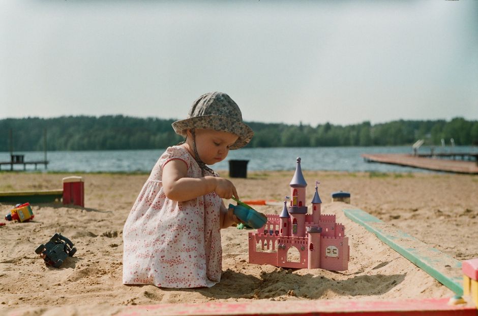 A young girl in a bucket hat plays with a toy castle on a sandy beach by the water.