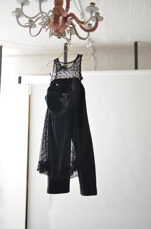 Elegant transparent dress and black trousers hanging on hanger placed on wooden chandelier in light room in daylight