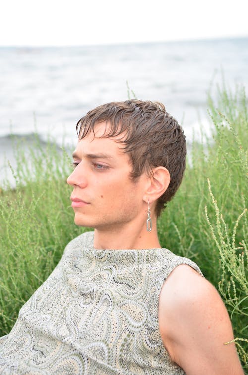 Thoughtful dark haired male with earring in trendy outfit relaxing on grass while looking away