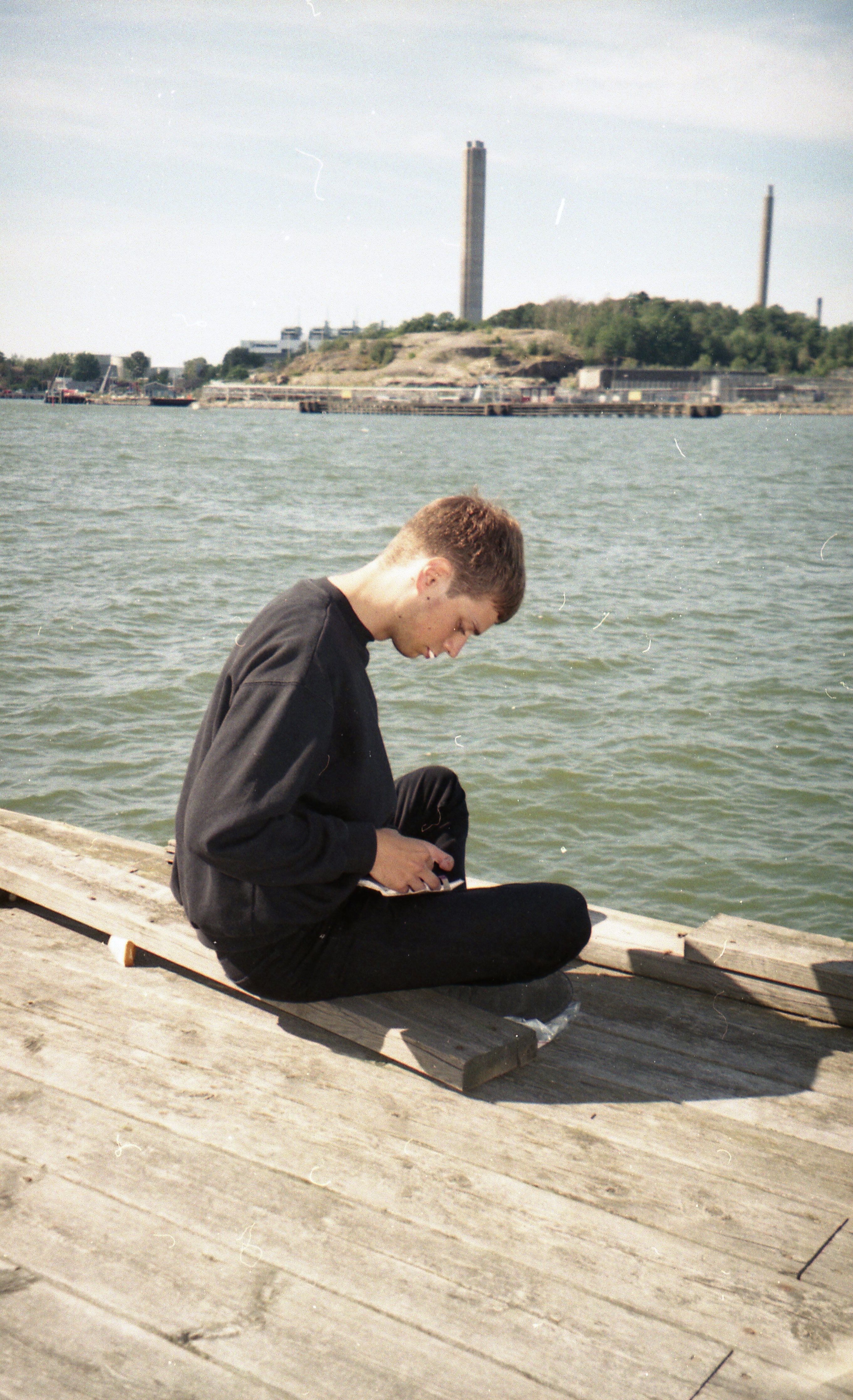 Side view of stylish male in casual outfit sitting with crossed legs on wooden embankment near rippling river