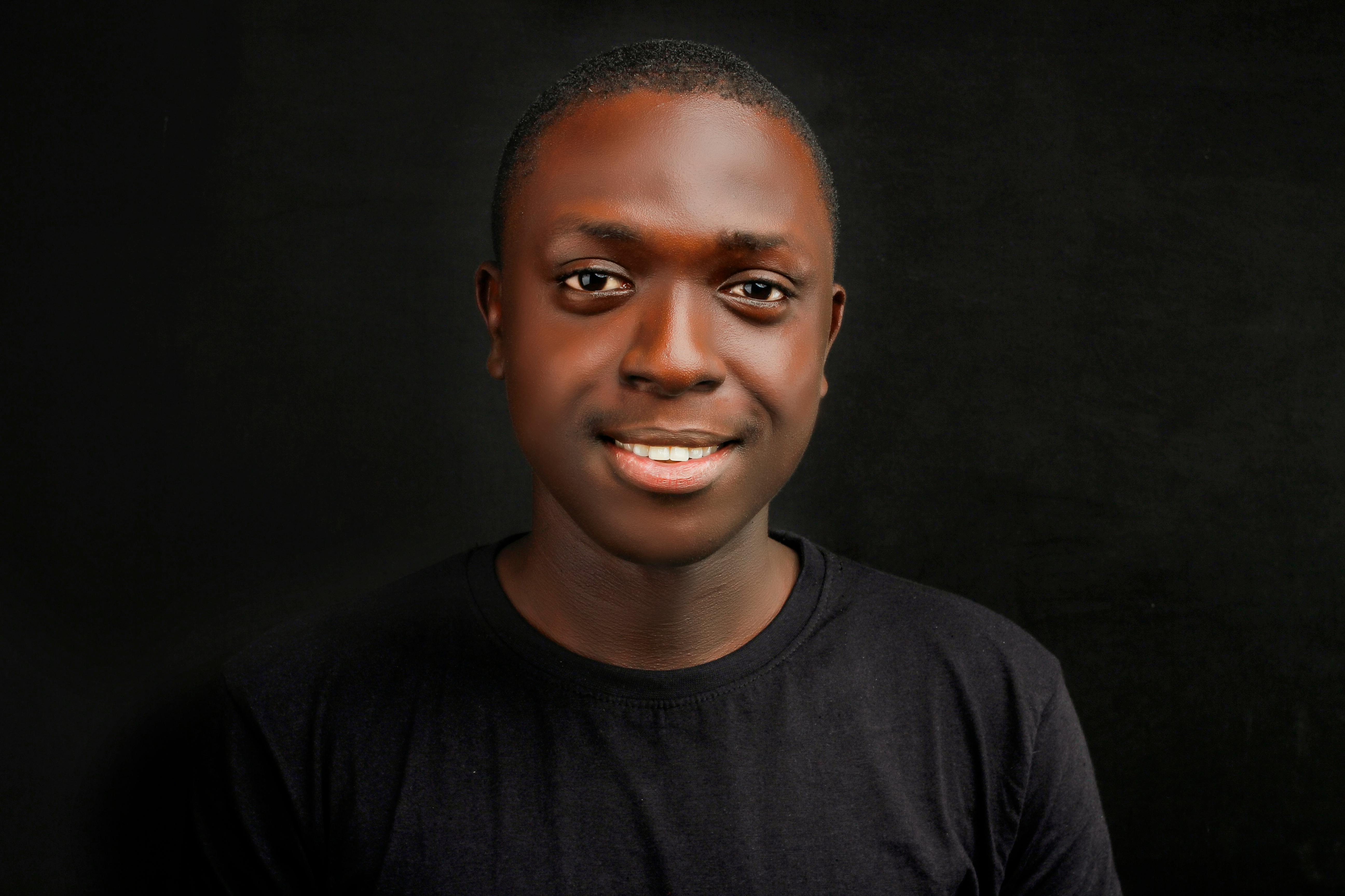 Close-up portrait of a smiling young African man in a black shirt against a dark background.