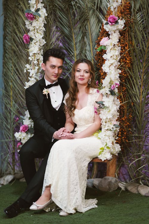 A Man in Blck Suit and Woman in White Gown Sitting on the Swing with Floral Decorations