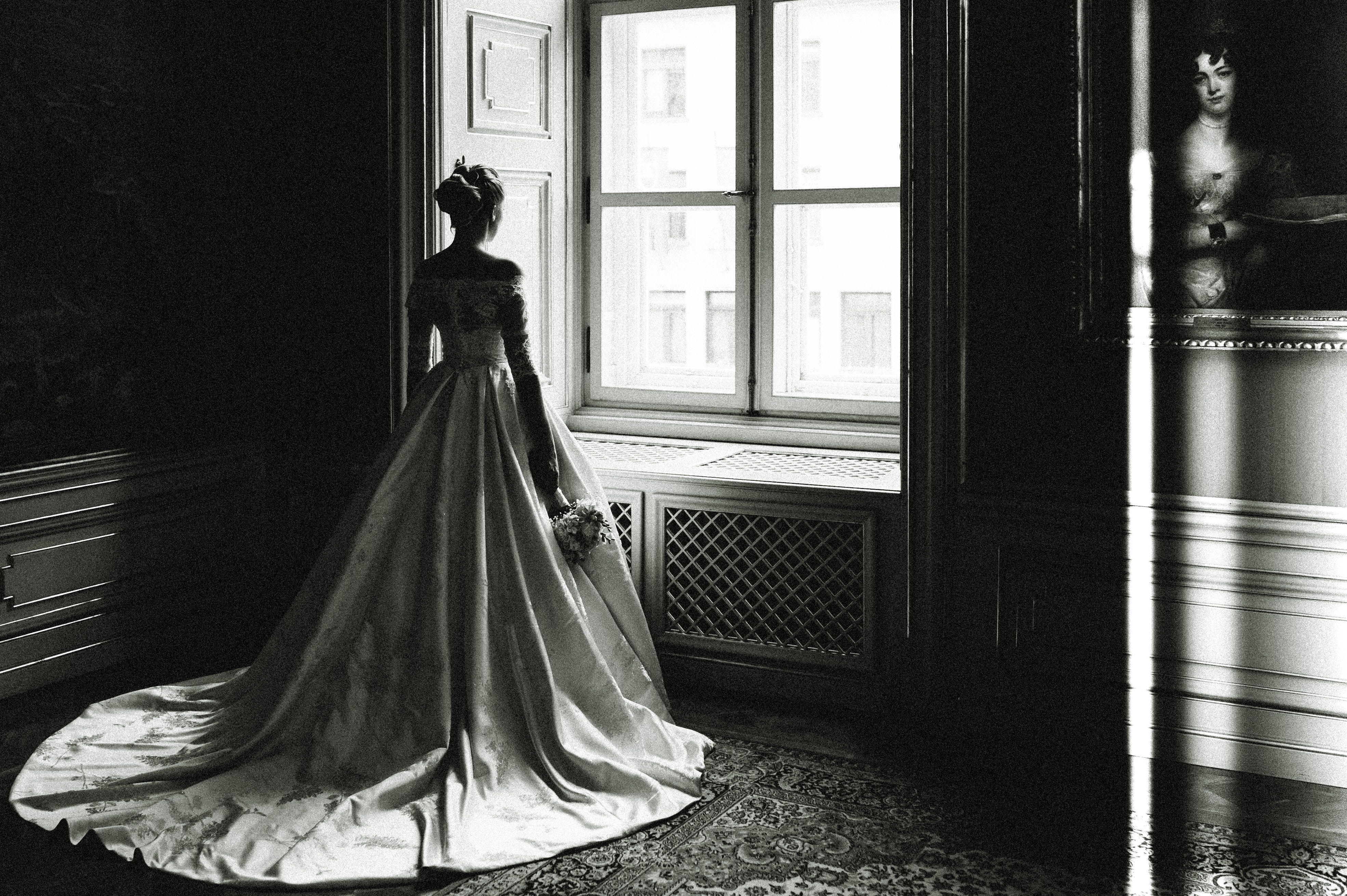 A dramatic grayscale image of a bride in a wedding gown standing by a window inside a classic room.