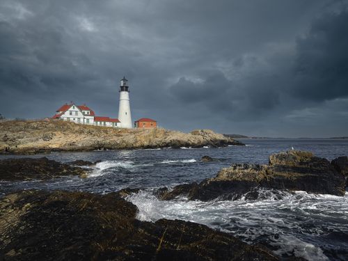 Free Lighthouse and houses located on shore with rocks washed by foamy waves in gloomy overcast day Stock Photo