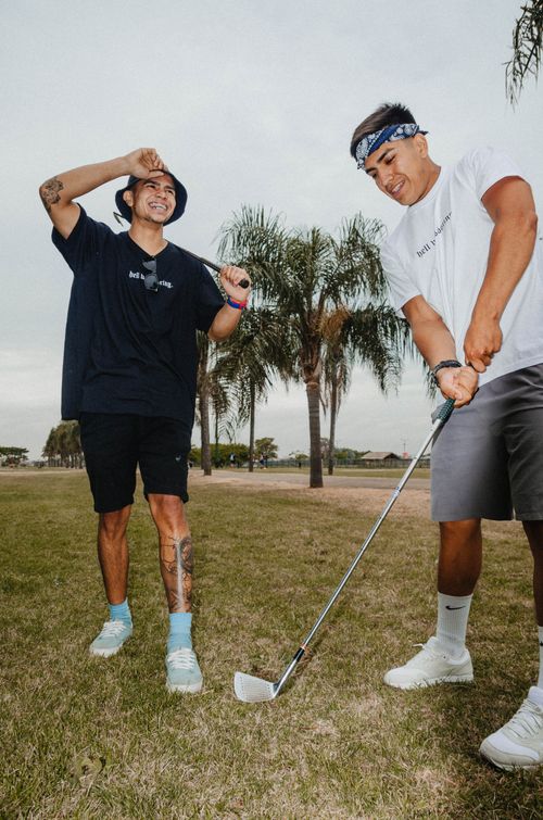 Free Two young men playing golf, smiling and enjoying a fun day outdoors on the green grass with palm trees behind. Stock Photo