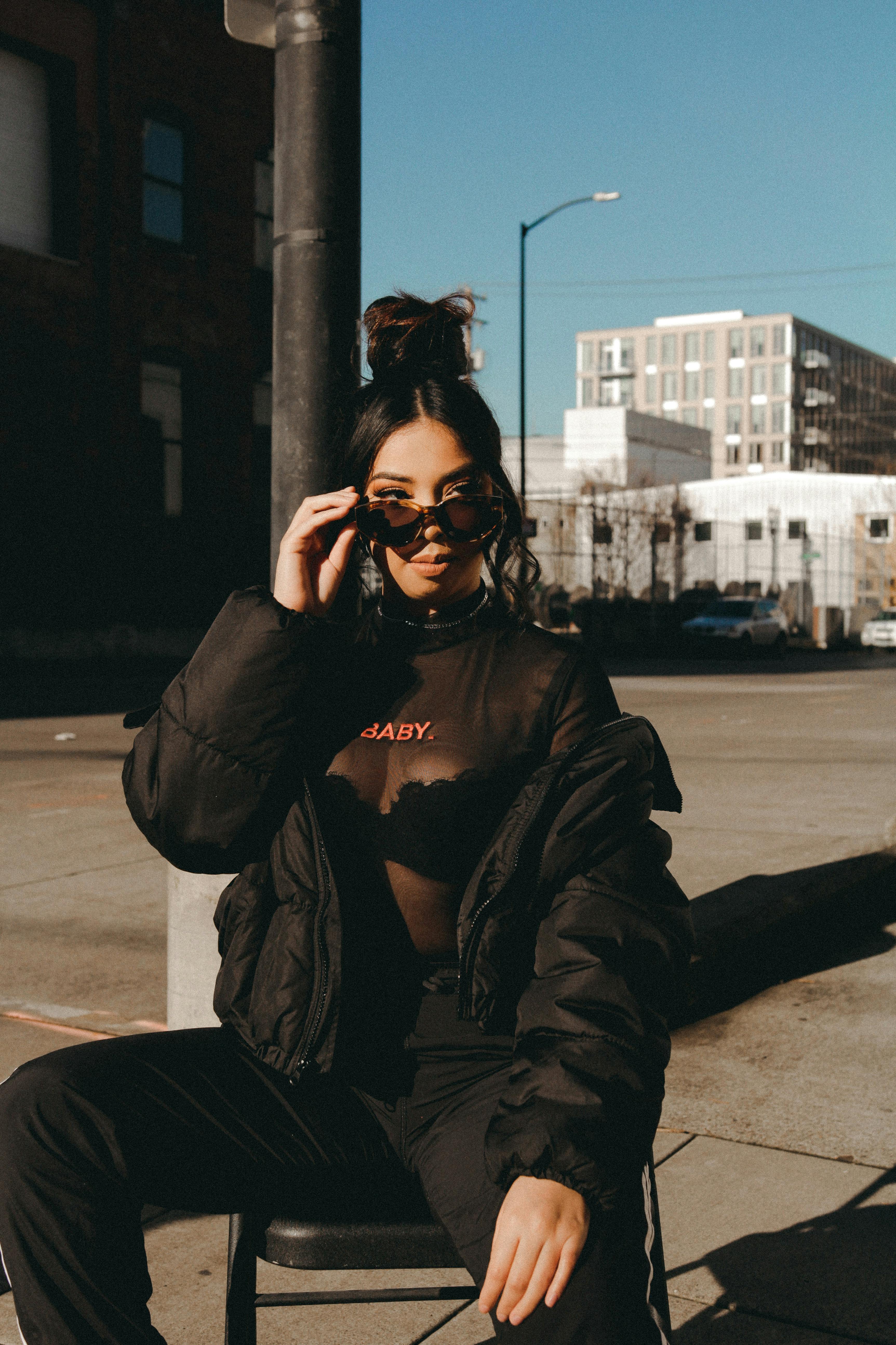 stylish young ethnic woman adjusting sunglasses while sitting on chair on street