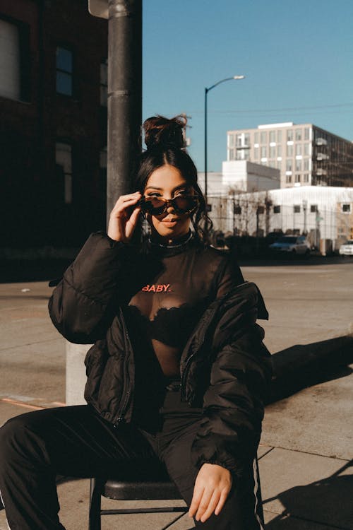 Free Self assured young ethnic female millennial in trendy outfit sitting on chair and adjusting sunglasses while chilling on city street on sunny day Stock Photo