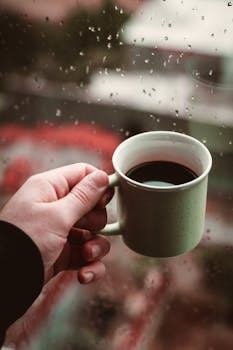 A hand holding a coffee cup by a rainy window, offering a cozy and warm vibe.
