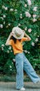Woman in Yellow Shirt and Blue Denim Jeans Wearing White Hat Standing Beside Pink Flowers