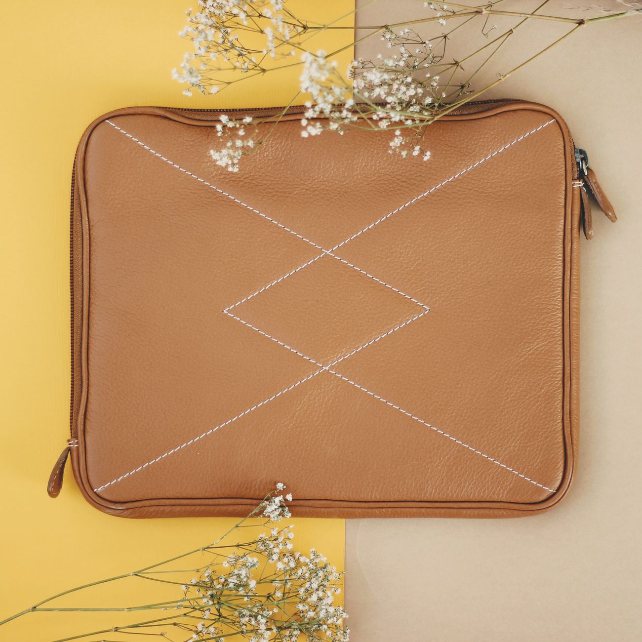 Top view of a stylish leather bag with stitching details on a pastel backdrop, decorated with white flowers.