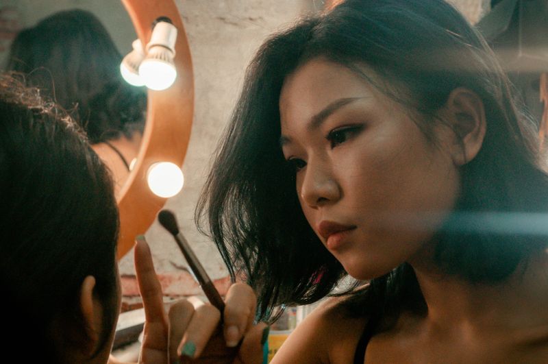 Asian woman using a makeup brush, applying makeup in front of a mirror