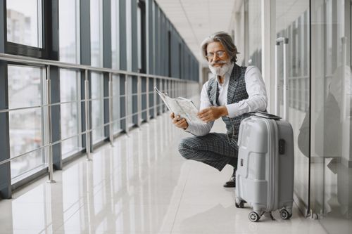Free Senior man in airport hallway holding a map, sitting beside luggage, illustrating travel and exploration. Stock Photo