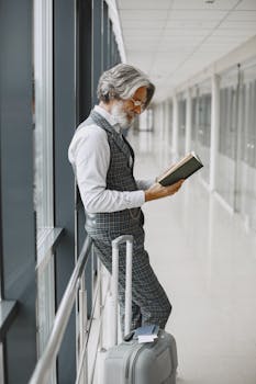 A mature man with gray hair reading a book in an airport hallway, leaning on luggage.