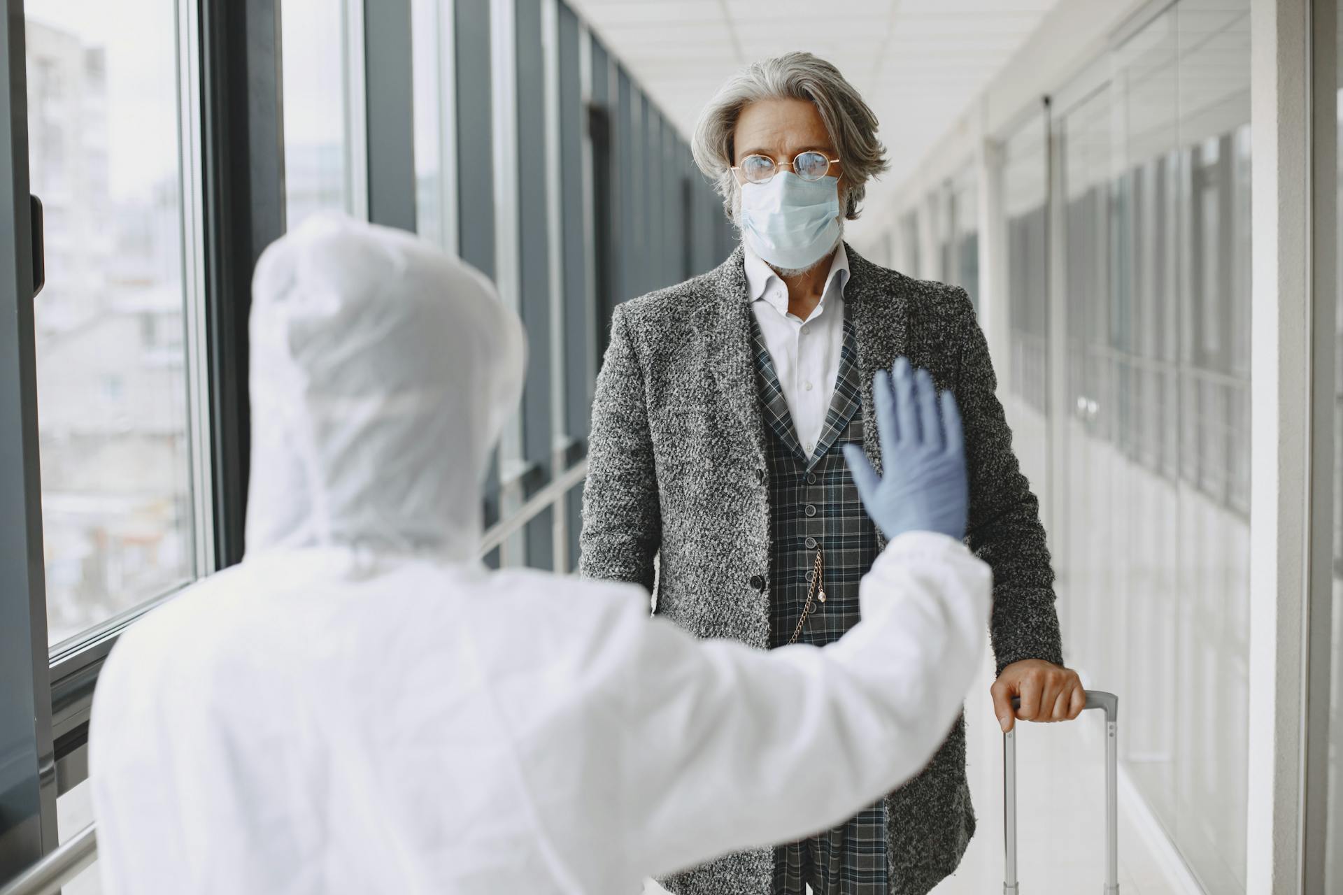 Traveler in face mask interacts with PPE-suited official in airport during pandemic inspection.