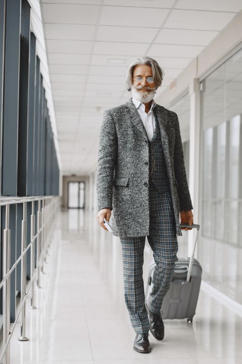 Free Senior man with gray hair and beard walking through airport hallway with luggage, wearing a gray blazer. Stock Photo