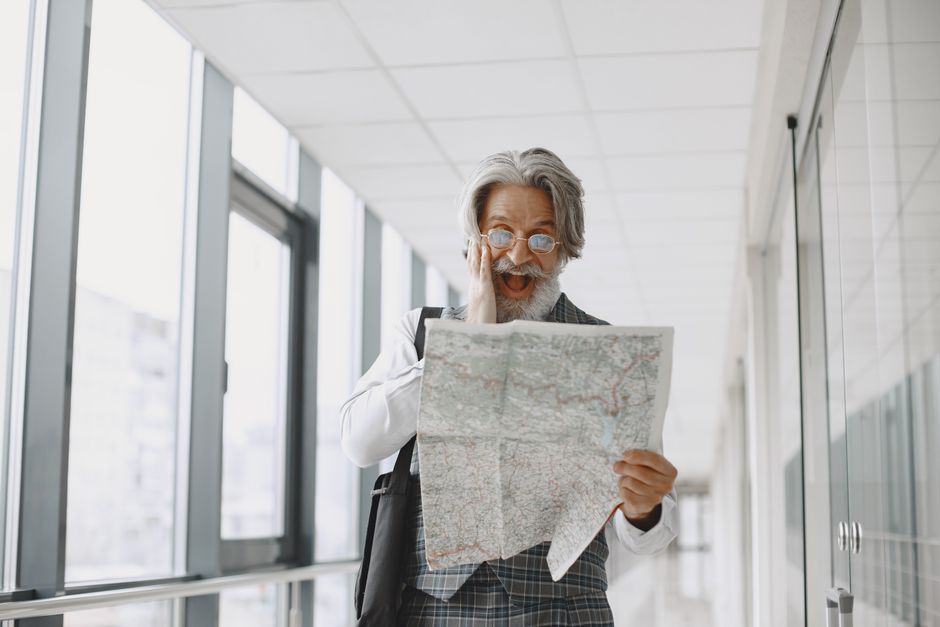 Elderly man with gray hair and eyeglasses looking surprised while holding a map indoors. Elderly man with gray hair and eyeglasses looking surprised while holding a map indoors.