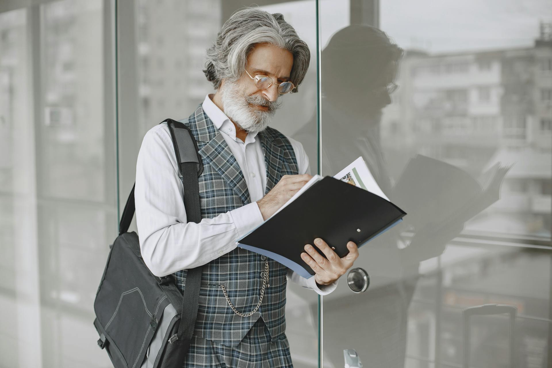 Elderly man in stylish office attire reading documents by glass windows.