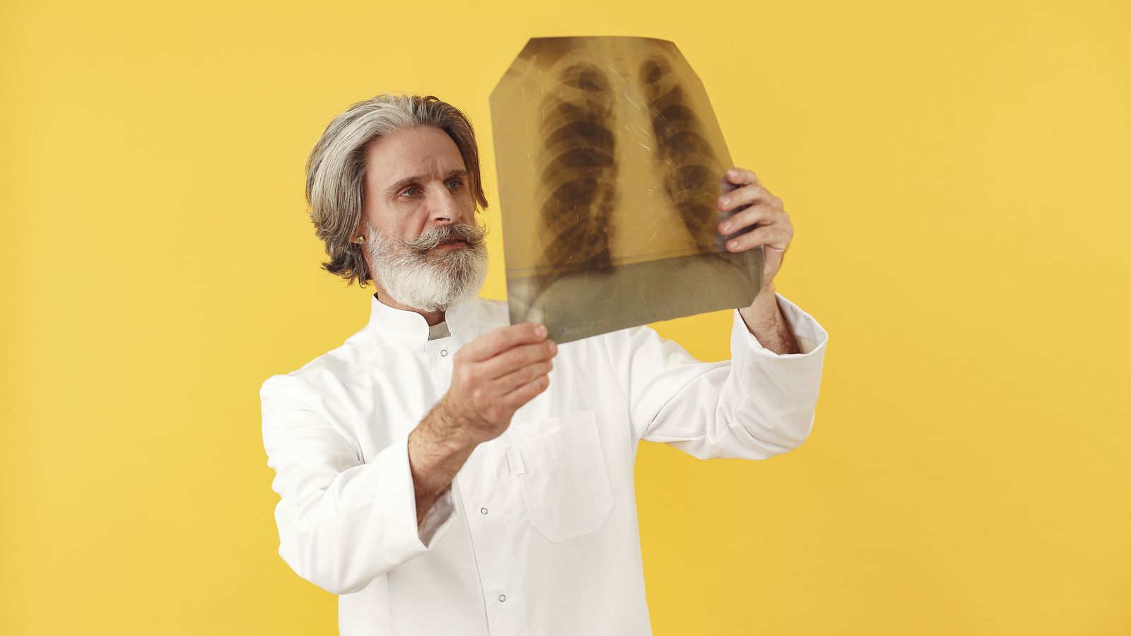 Senior male doctor holding and examining a chest x-ray, posing against a bright yellow studio background.
