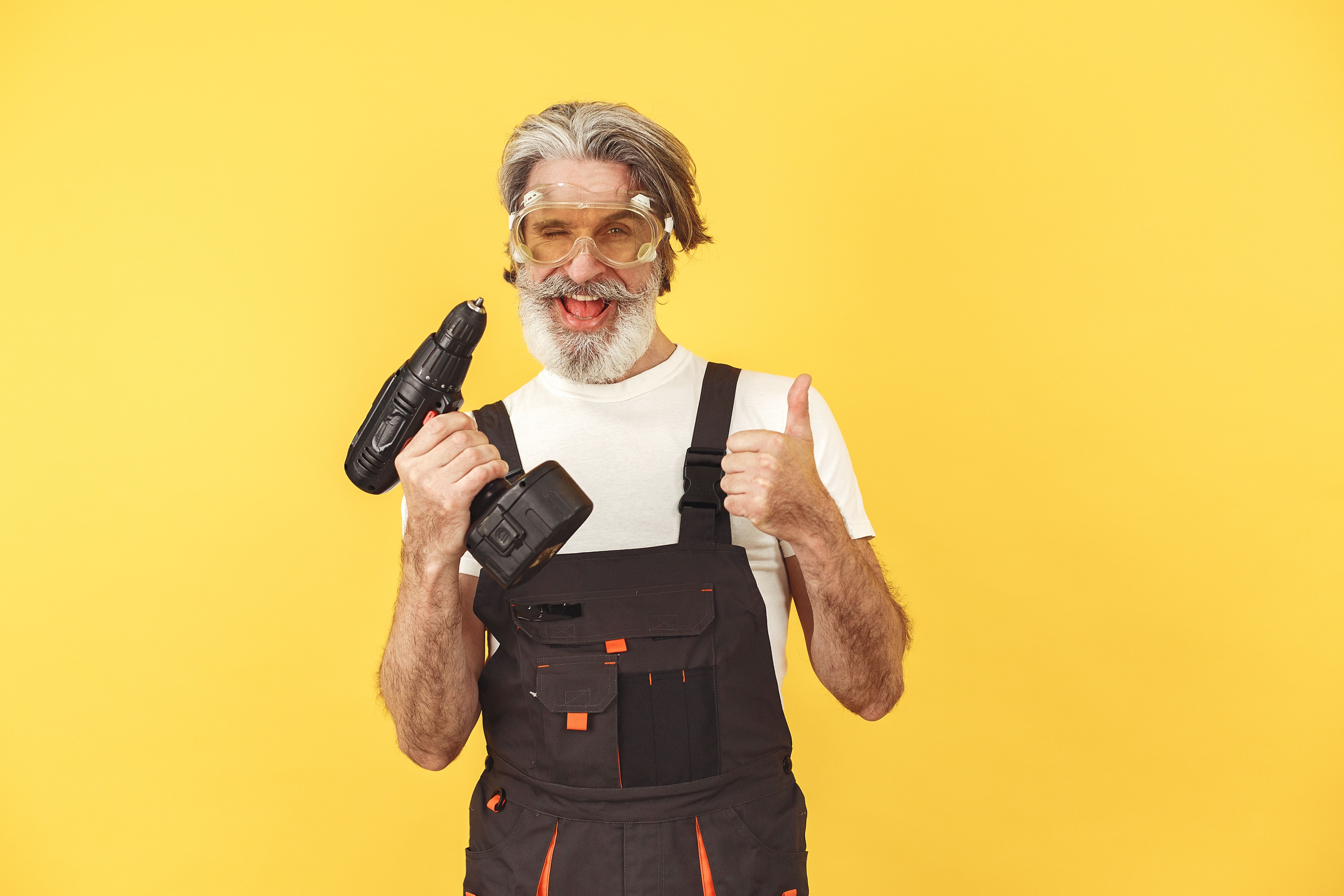 Senior man in safety gear and overalls holding a drill, smiling against a yellow background.