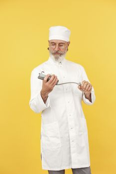Elderly male doctor in white coat and cap examining medical tool against a vibrant yellow background.