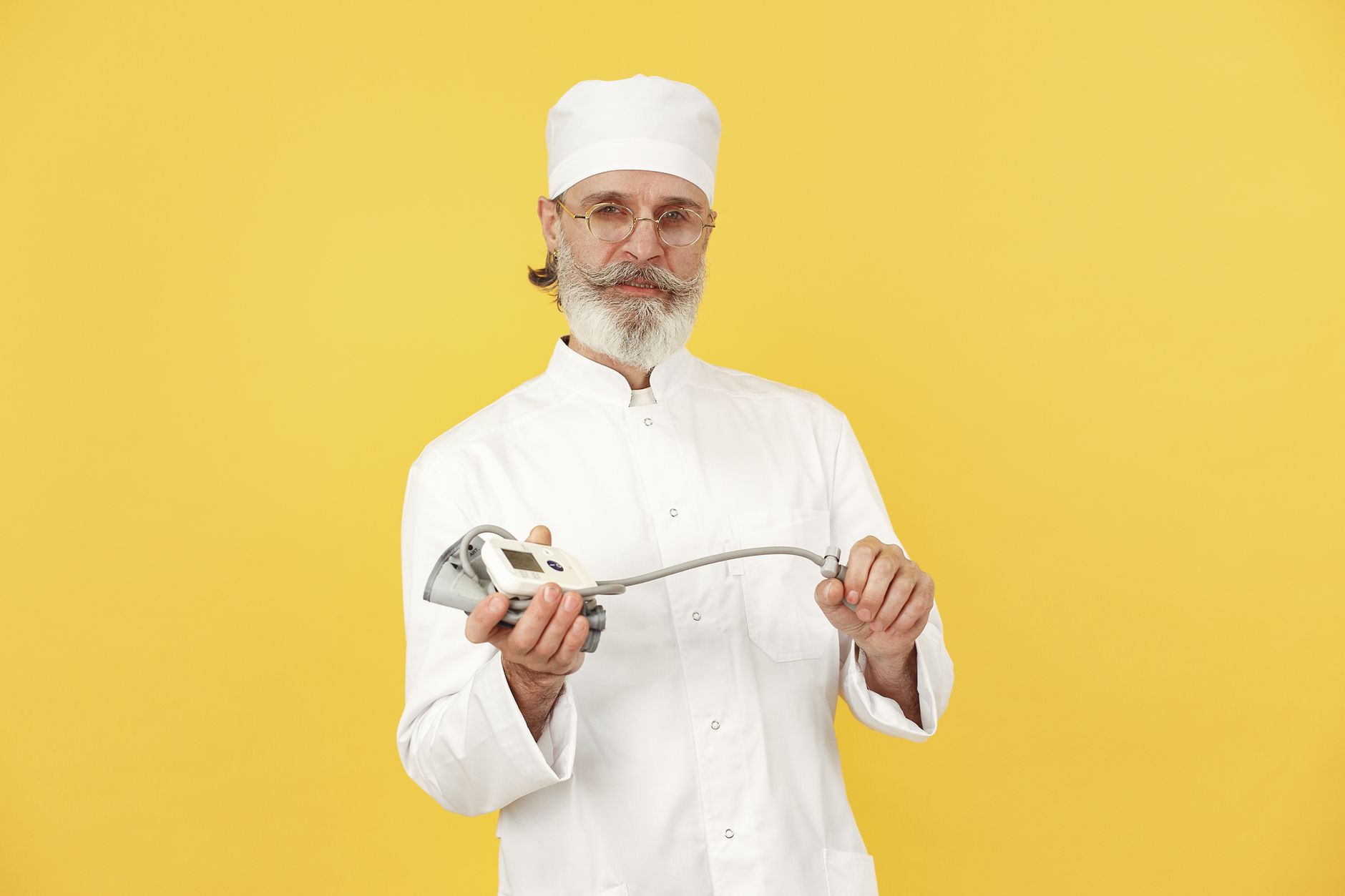 A senior chef in white attire holding a kitchen blender, standing against a yellow background.