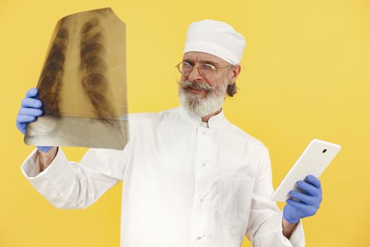 Senior male doctor examines an X-ray while holding a tablet computer against a yellow studio background.