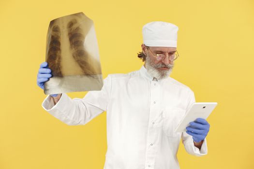 Bearded senior doctor in white coat examining an x-ray while holding a tablet against a yellow background.