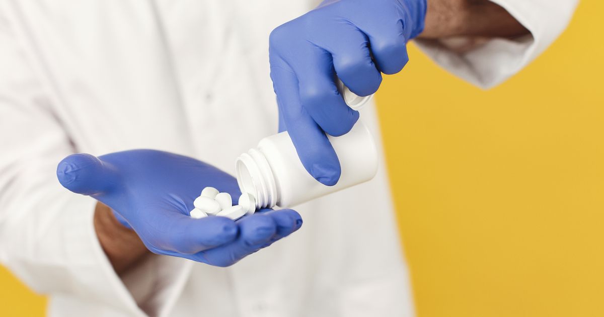 Pharmacist in a white coat wearing blue gloves pouring white pills from a bottle into a hand