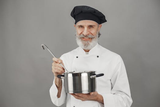 A cheerful senior chef with a beard holds a cooking pot and ladle, wearing a chef hat.