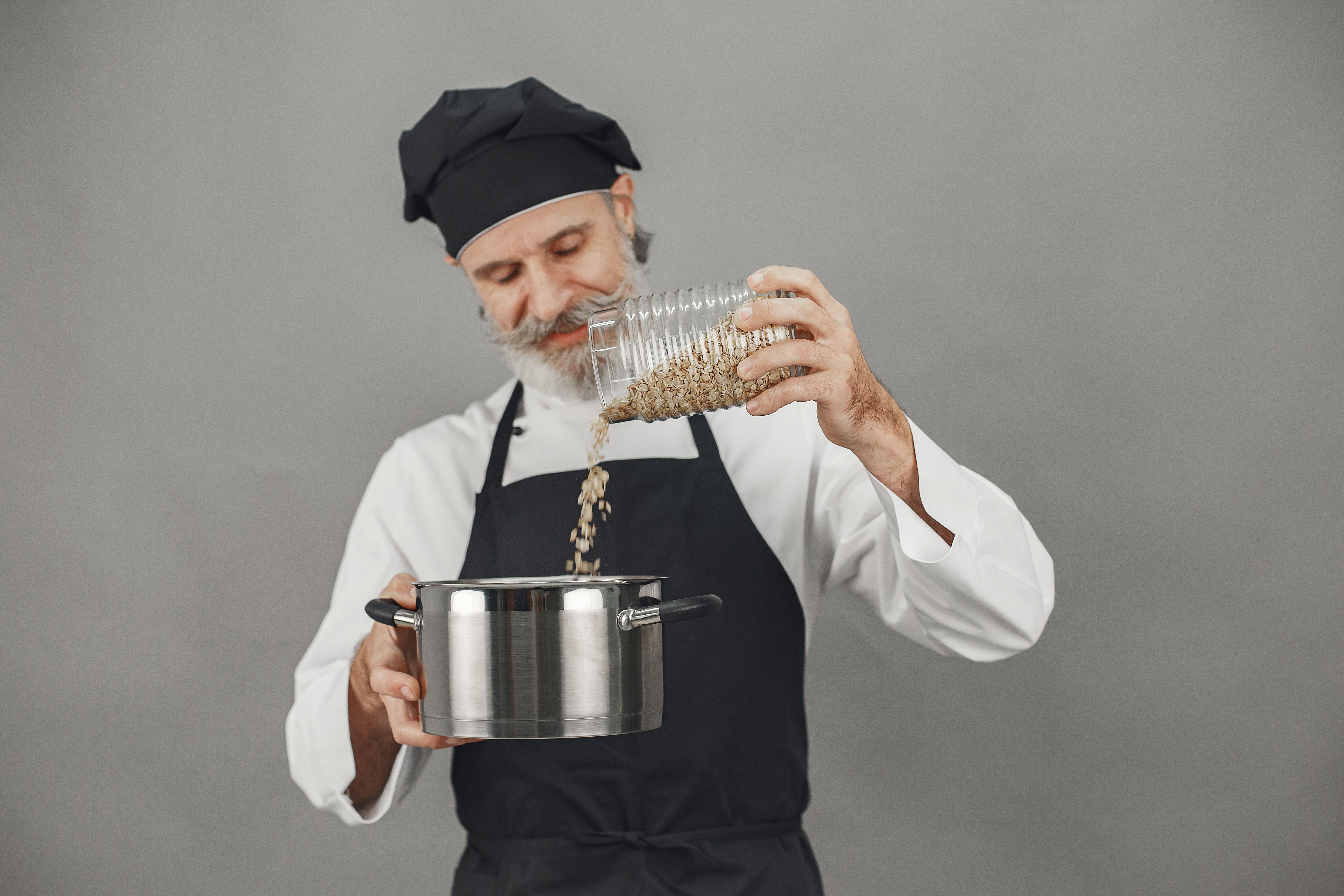An experienced chef in uniform pours oats into a pot, focusing on culinary precision.