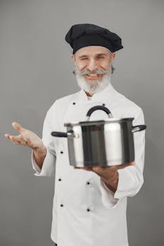 A cheerful senior chef in uniform presenting a cooking pot, showcasing culinary expertise.