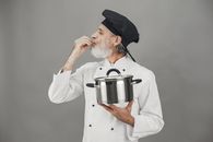 Chef in uniform tasting dish with cooking pot, making hand gesture of satisfaction.