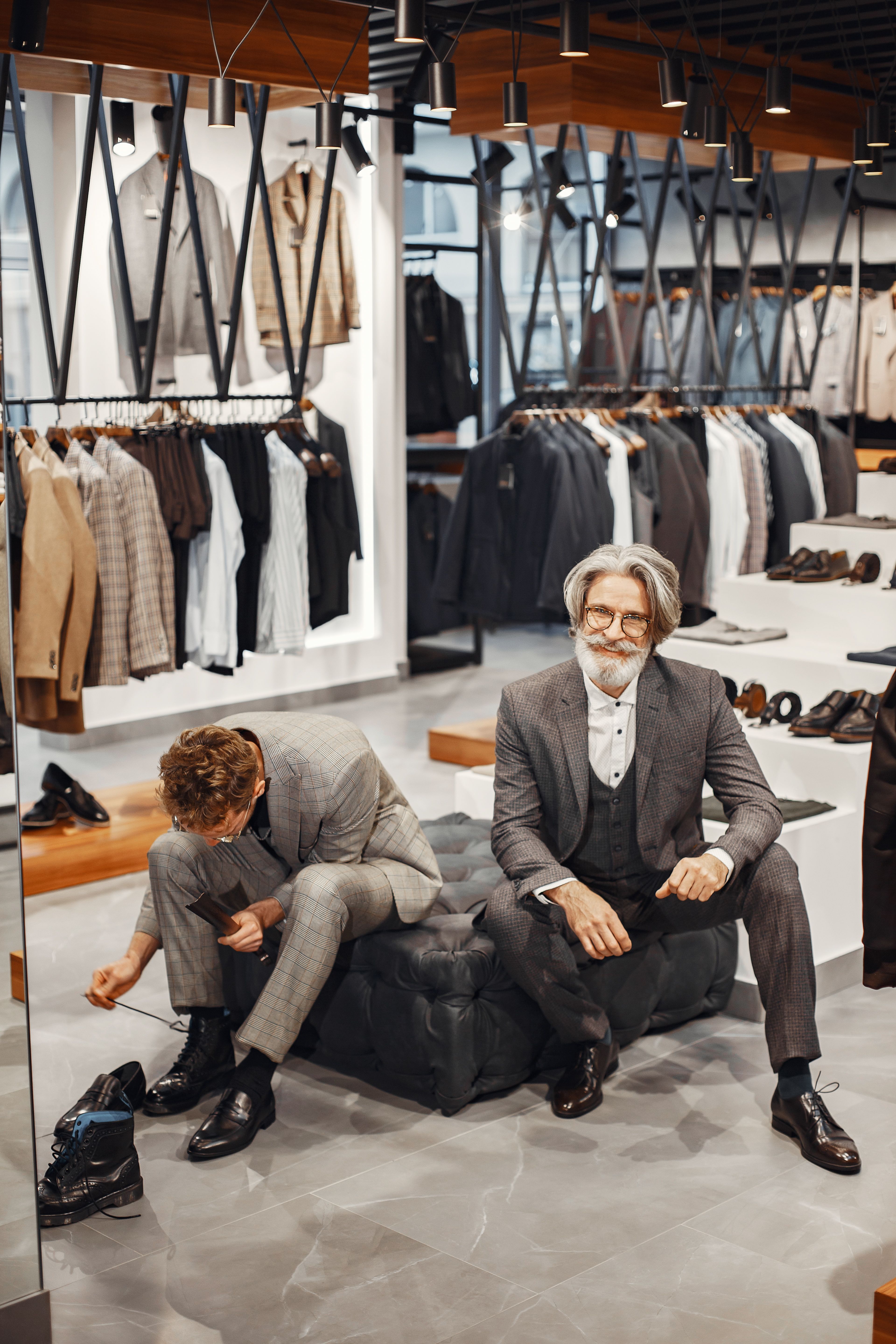 Two men wearing formal suits in a stylish men's suit shop with a variety of clothing on display.