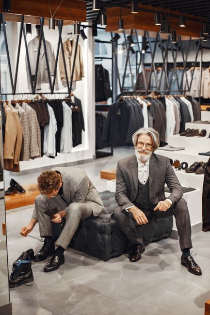 Two men wearing formal suits in a stylish men's suit shop with a variety of clothing on display. Two men wearing formal suits in a stylish men's suit shop with a variety of clothing on display.