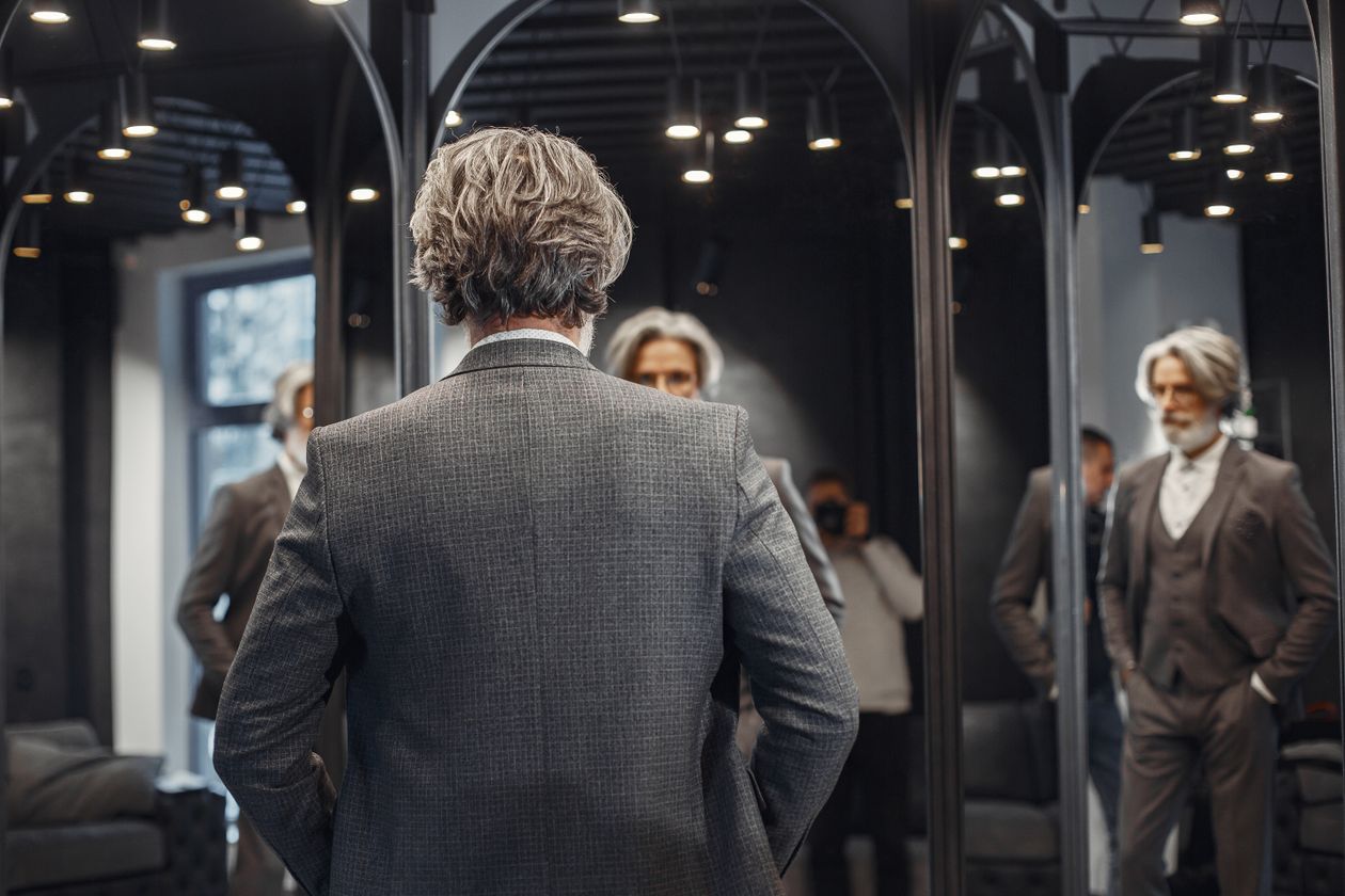 A man in a grey suit looking at his reflection in a shop mirror