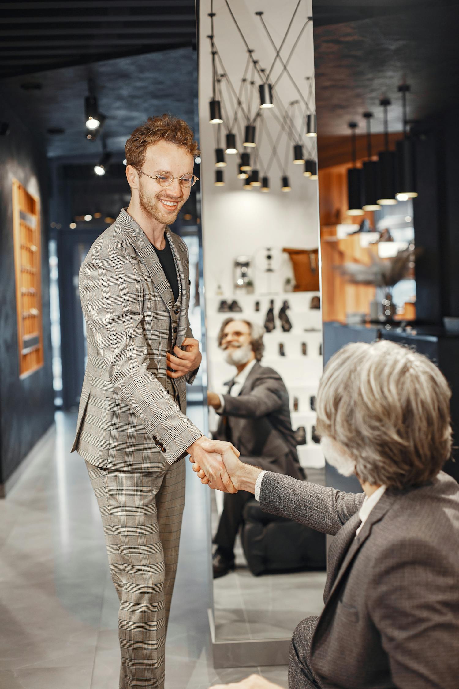 Two caucasian men in elegant suits shaking hands in a modern office setting, smiling.