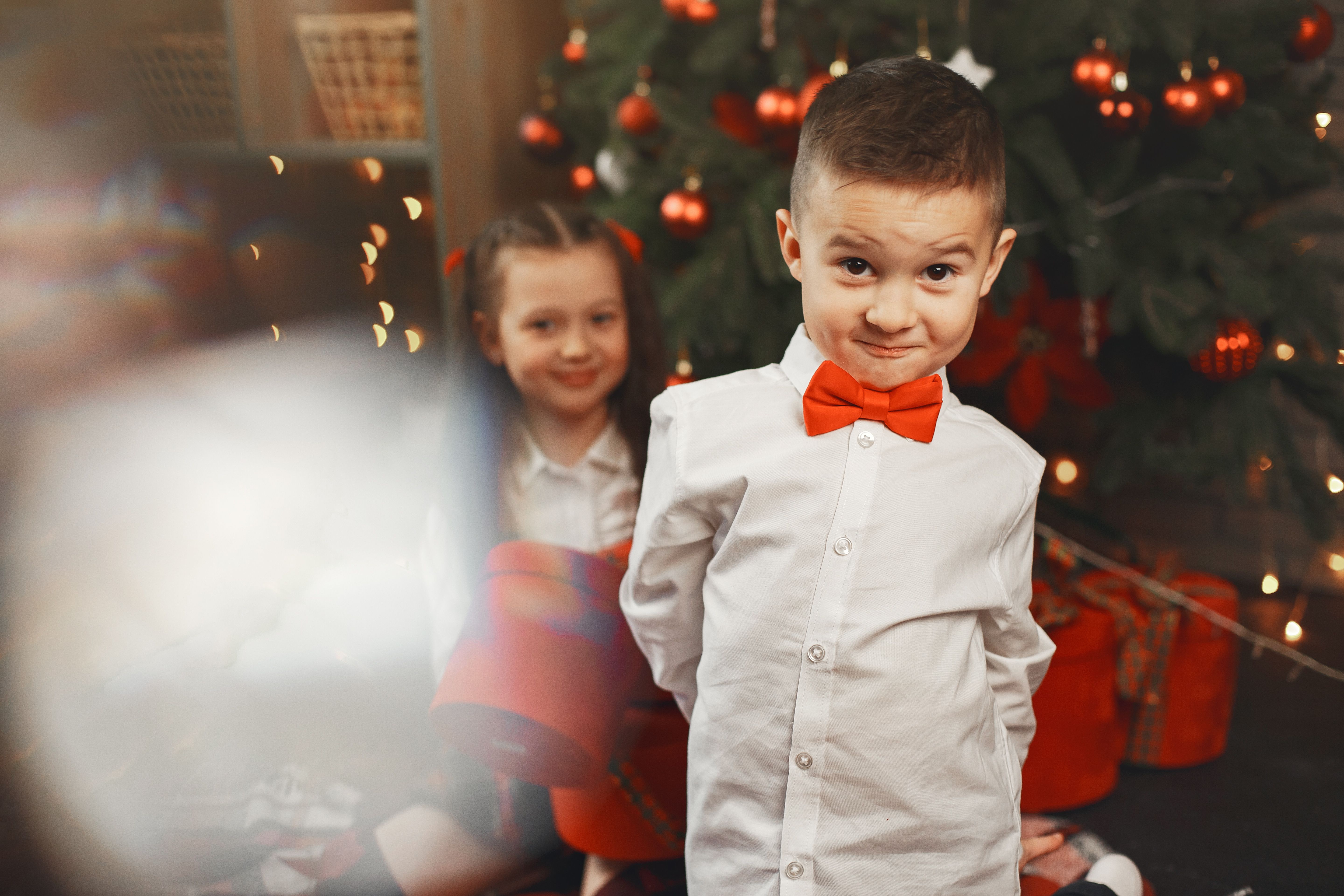 Two children in white shirts and red accents celebrating beside a Christmas tree with gifts.
