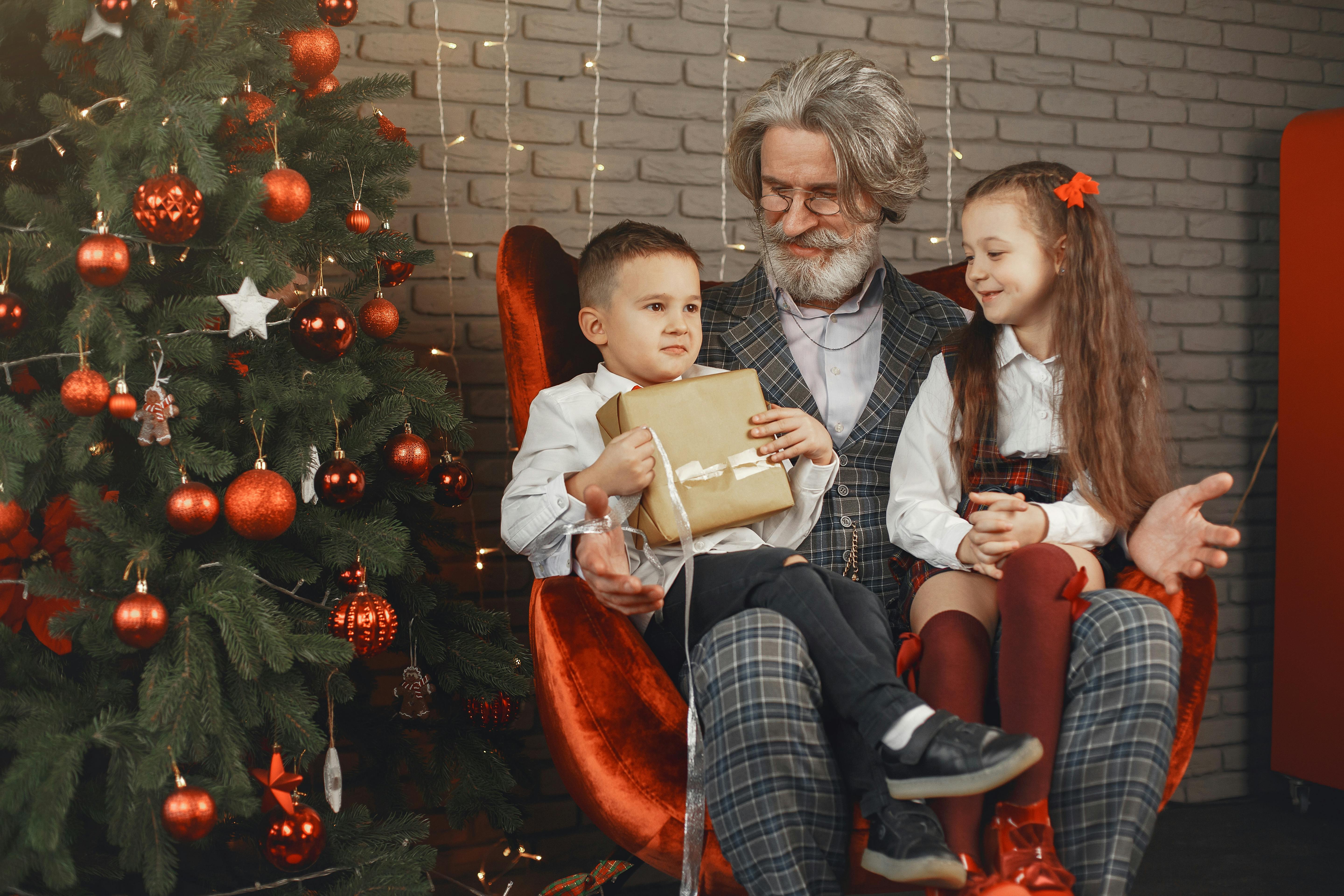 Grandfather and grandchildren celebrating Christmas, sitting by the tree with presents and joyful expressions.