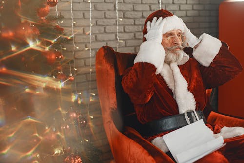 Man Dressed As Santa Claus Sitting Next to a Christmas Tree and Holding His Head